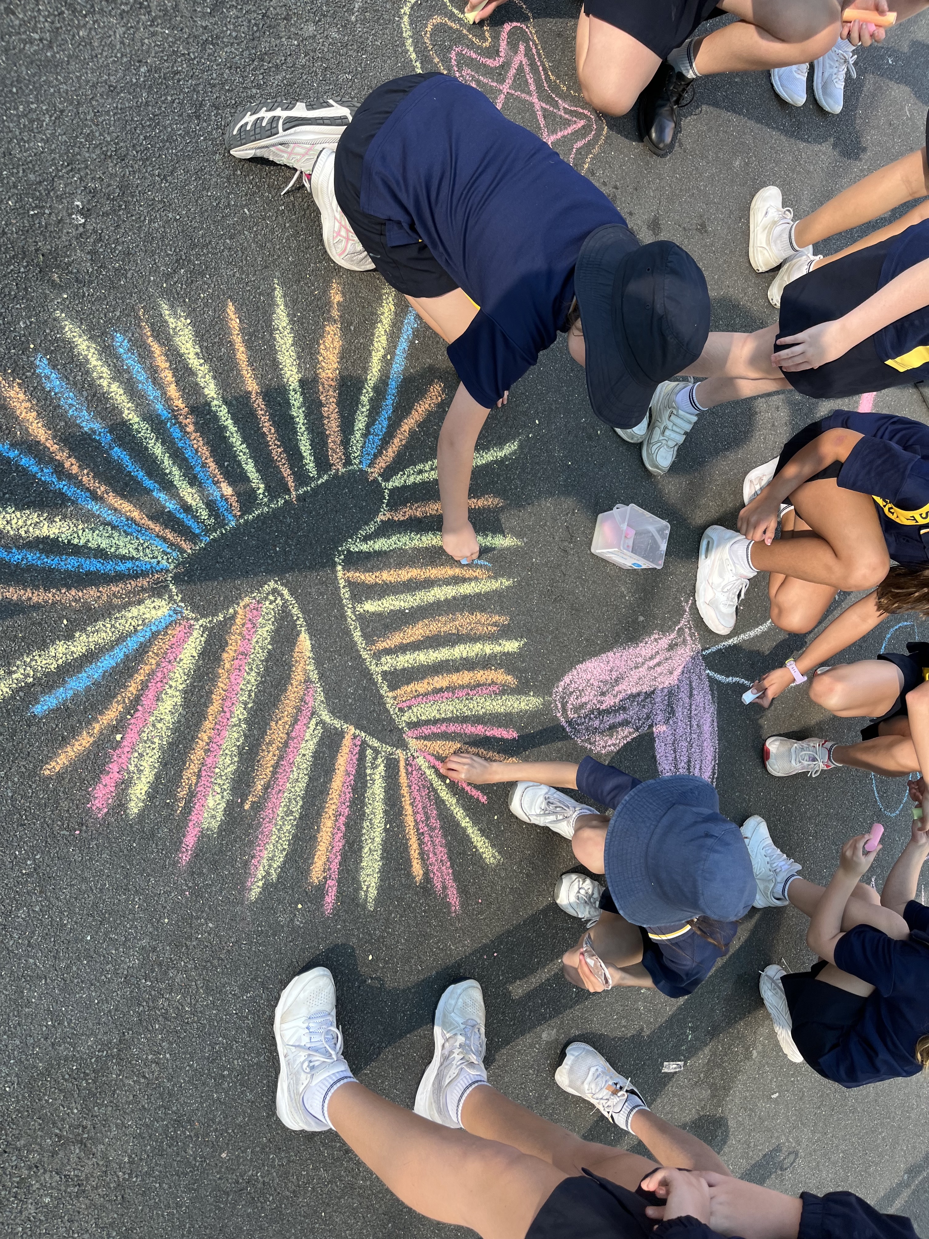 Students sit and kneel in a circle on an asphalt playground, drawing a large tau cross with colourful chalk. Radiating lines in pink, yellow, blue and green extend evenly from the centre, creating a bright, symmetric design as hands add the final details.